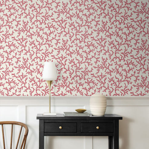 A styled interior corner showing a black console table against a white wainscoted wall with red coral-pattern wallpaper. A white table lamp with a gold stand, a ribbed white ceramic vase, and a small gold bowl sit on the table, with a wooden chair partially visible on the left.
