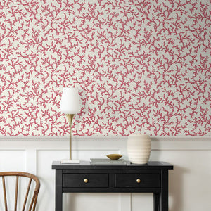 A styled interior corner showing a black console table against a white wainscoted wall with red coral-pattern wallpaper. A white table lamp with a gold stand, a ribbed white ceramic vase, and a small gold bowl sit on the table, with a wooden chair partially visible on the left.