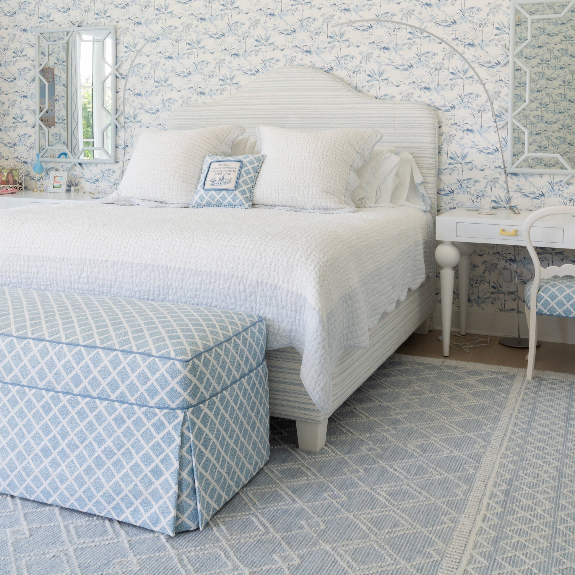 A beautifully styled bedroom featuring a white bed with white linens, a light blue and white patterned upholstered ottoman, a matching patterned chair, and a light blue and white geometric rug. The wall behind the bed is covered in matching blue and white floral toile wallpaper.