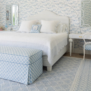 A beautifully styled bedroom featuring a white bed with white linens, a light blue and white patterned upholstered ottoman, a matching patterned chair, and a light blue and white geometric rug. The wall behind the bed is covered in matching blue and white floral toile wallpaper.