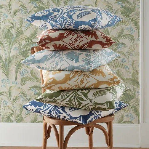 Stack of colorful patterned pillows on a wooden stool against a floral wallpapered wall.