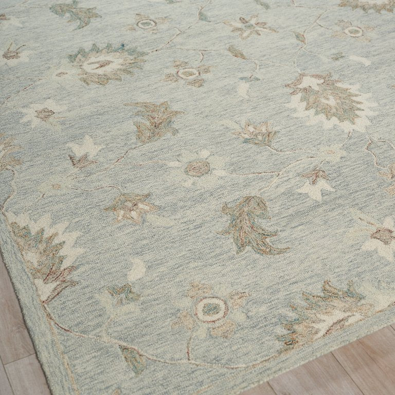 A close-up view of a light blue-gray area rug with a distressed floral and botanical pattern in beige, cream, and muted sage green, shown on a light wood floor.