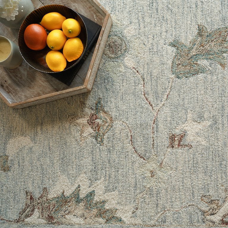 A top-down view of a textured area rug with a muted blue-gray base and a floral vine pattern in beige, brown, and soft green. A dark wooden tray holding oranges and a candle sits on the rug in the upper left corner.