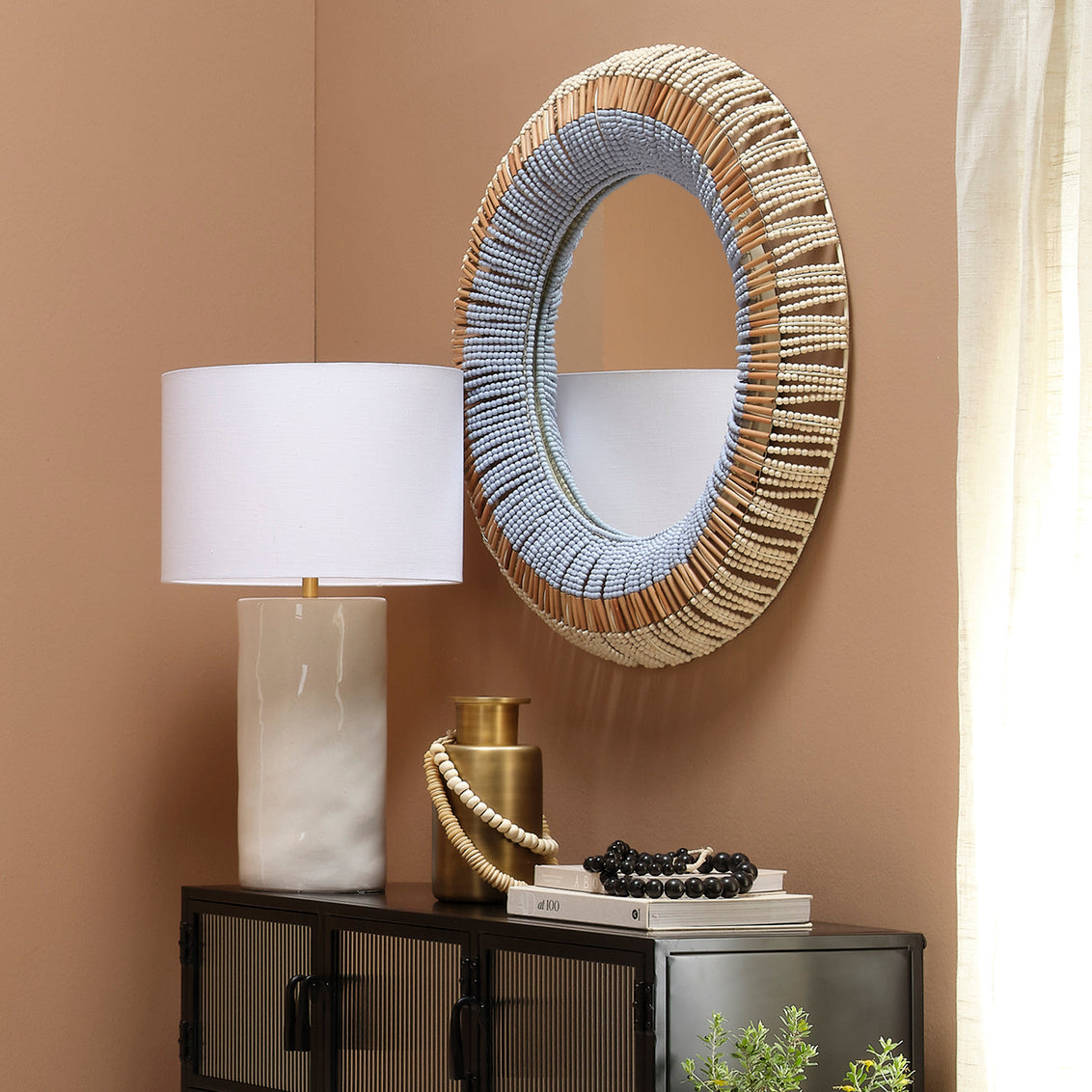 Decorative round mirror with a textured frame on a beige wall above a dark cabinet with a lamp and decorative items.