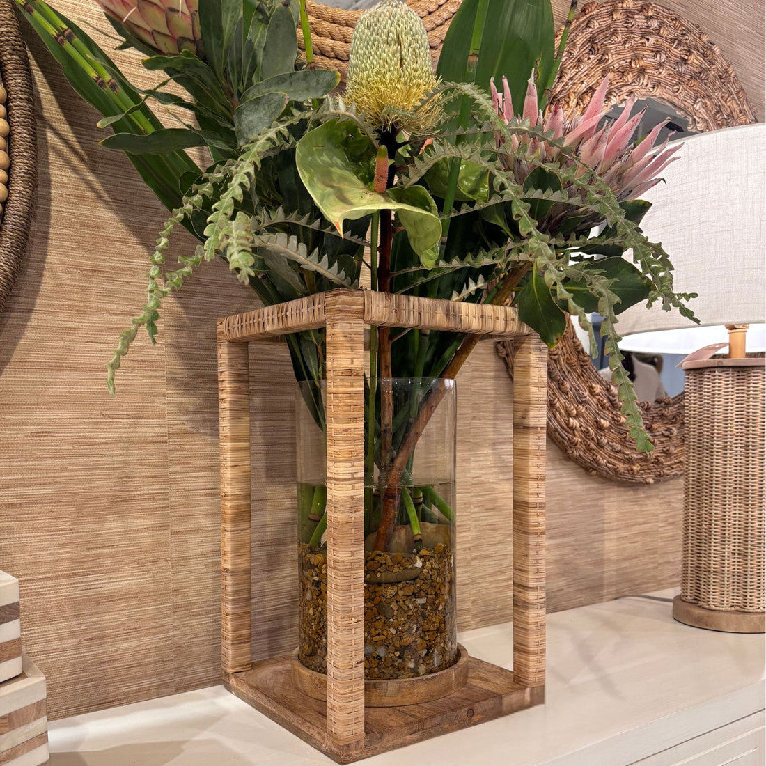 Decorative vase with plants on a wooden stand against a wicker wall.