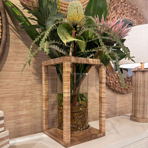 Decorative vase with plants on a wooden stand against a wicker wall.