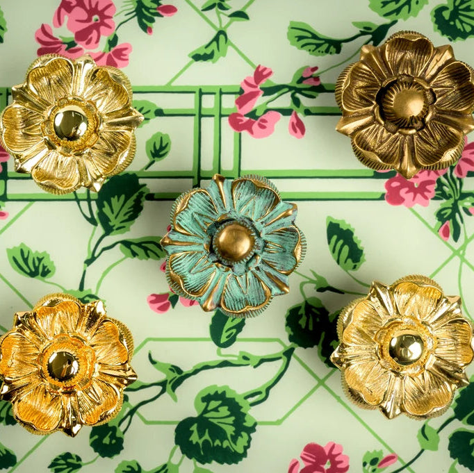 Five ornate flower-shaped decorative knobs arranged on a light green floral wallpaper background. Four knobs are metallic gold, and the center knob has a distressed turquoise-blue finish with a gold center.