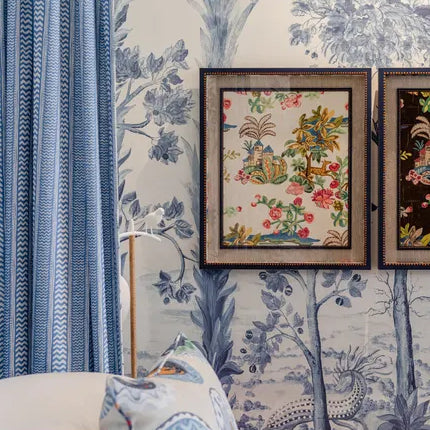 Two framed decorative art prints with colorful floral and bird motifs, mounted on a blue and white toile wallpapered wall next to a blue curtain.
