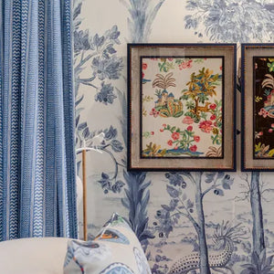 Two framed decorative art prints with colorful floral and bird motifs, mounted on a blue and white toile wallpapered wall next to a blue curtain.