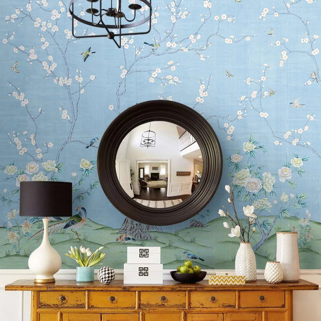 A styled interior entryway featuring a light blue floral wallpaper with white blossoms and birds, a round black-framed mirror, a yellow wooden console table, a white table lamp, and decorative vases.