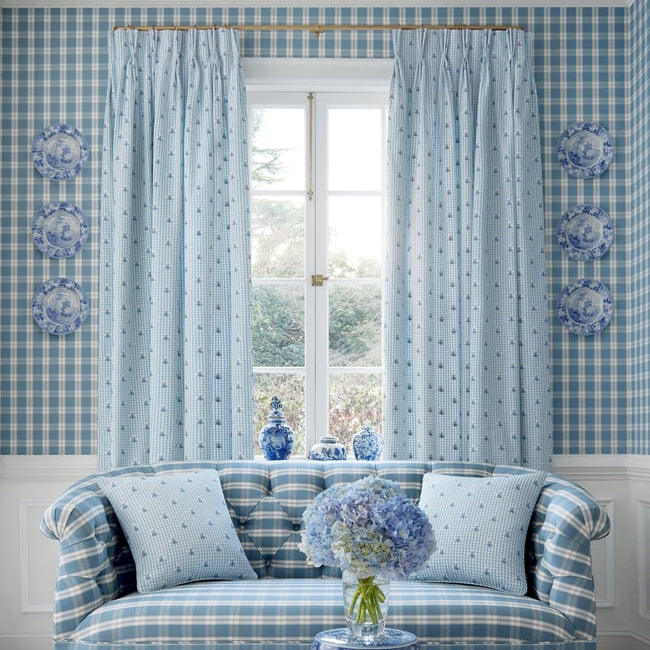 A styled interior scene featuring a blue and white gingham sofa with matching pillows, light blue gingham curtains, blue-and-white patterned wallpaper, and a blue-and-white ceramic stool in the foreground.