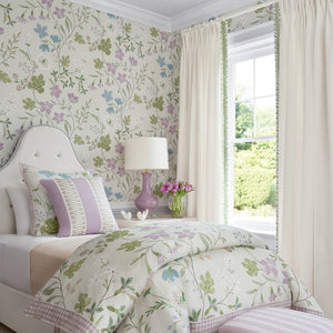 A beautifully styled bedroom featuring a bed dressed in a light floral print comforter and matching pillows, set against floral wallpaper. A rustic wooden stool with a pink and white striped cushion sits at the foot of the bed, and a window with cream curtains allows natural light into the room.