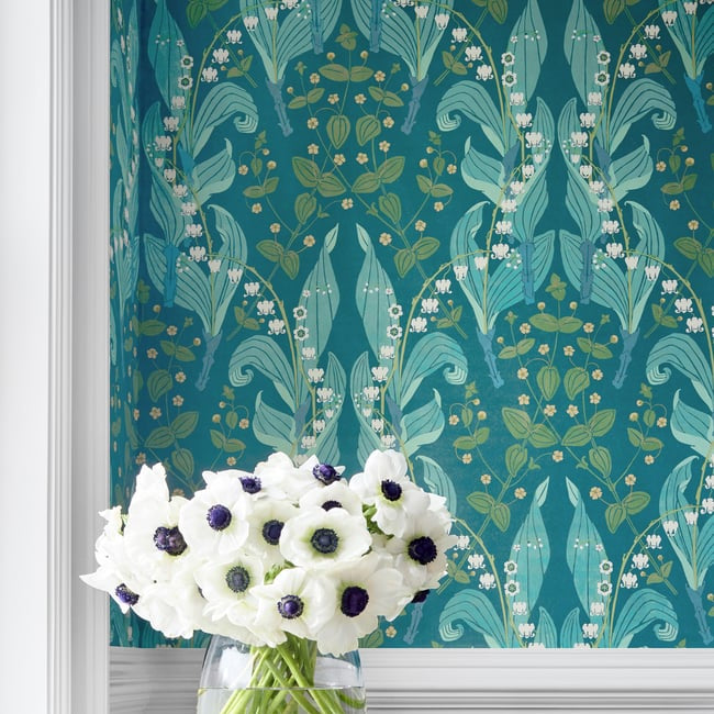 A styled interior scene showing a teal and aqua botanical wallpaper pattern on a wall above a light wood side table. The table holds a clear glass vase filled with white anemone flowers with dark centers, stacked on two blue books.