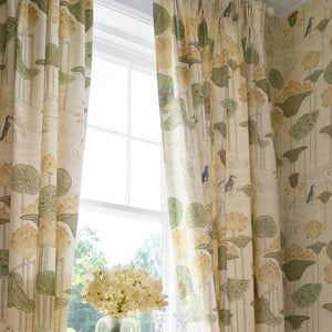 A window dressed with patterned curtains featuring a botanical print in green and beige, with matching wallpaper on the wall. A small woven console table with a vase of white flowers sits in front of the window.