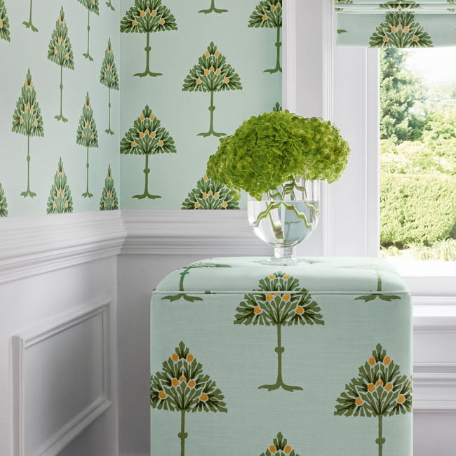 A styled interior corner showing wallpaper and a matching fabric ottoman or stool in a pale mint green color with a repeating tree pattern, next to a window with a matching roller blind and a green hydrangea arrangement.