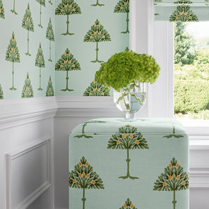 A styled interior corner showing wallpaper and a matching fabric ottoman or stool in a pale mint green color with a repeating tree pattern, next to a window with a matching roller blind and a green hydrangea arrangement.