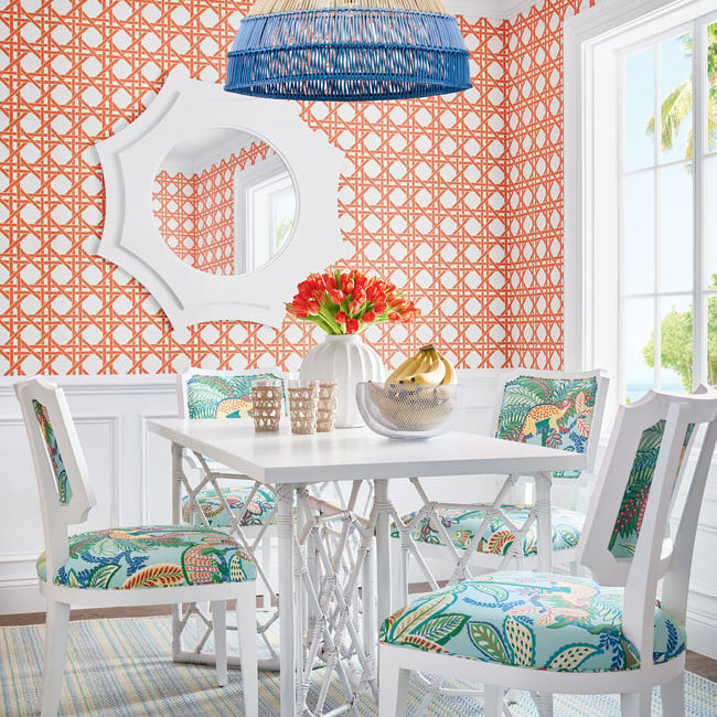 Dining room with white table and chairs with colorful cushions, orange geometric wallpaper, and a blue pendant light.