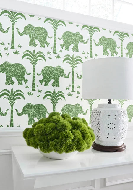 Decorative wall with green elephant and palm tree pattern, white table with green plant and lamp.