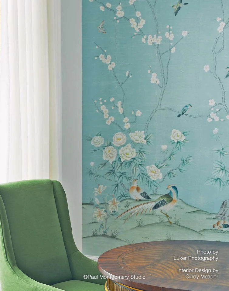 A styled interior corner featuring a large wall mural with a light blue-green background and delicate hand-painted style illustrations of white flowers, birds, and a pheasant. A green upholstered armchair and a round wooden table are partially visible in the foreground.
