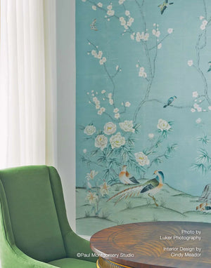 A styled interior corner featuring a large wall mural with a light blue-green background and delicate hand-painted style illustrations of white flowers, birds, and a pheasant. A green upholstered armchair and a round wooden table are partially visible in the foreground.
