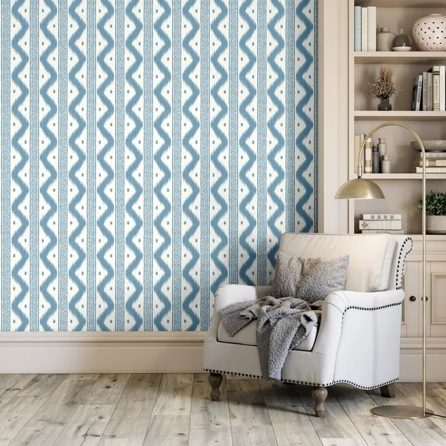 Blue and white patterned wallpaper shown in a styled room setting with a white armchair, gray throw blanket, gold floor lamp, and wooden bookshelves.