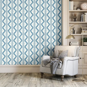 Blue and white patterned wallpaper shown in a styled room setting with a white armchair, gray throw blanket, gold floor lamp, and wooden bookshelves.