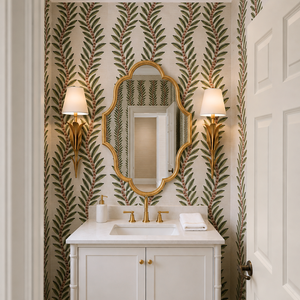 A white bathroom vanity with a white marble countertop, gold faucet, and an ornate gold-framed mirror, set against wallpaper with a green leaf pattern on a cream background. Two gold sconces with white shades flank the mirror.