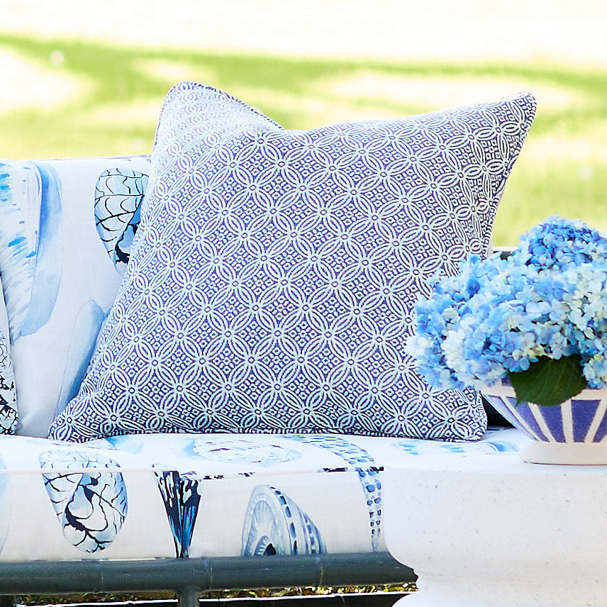 A blue and white patterned throw pillow and matching outdoor sofa cushions styled on a bench with a vase of blue flowers in a blurred garden background.