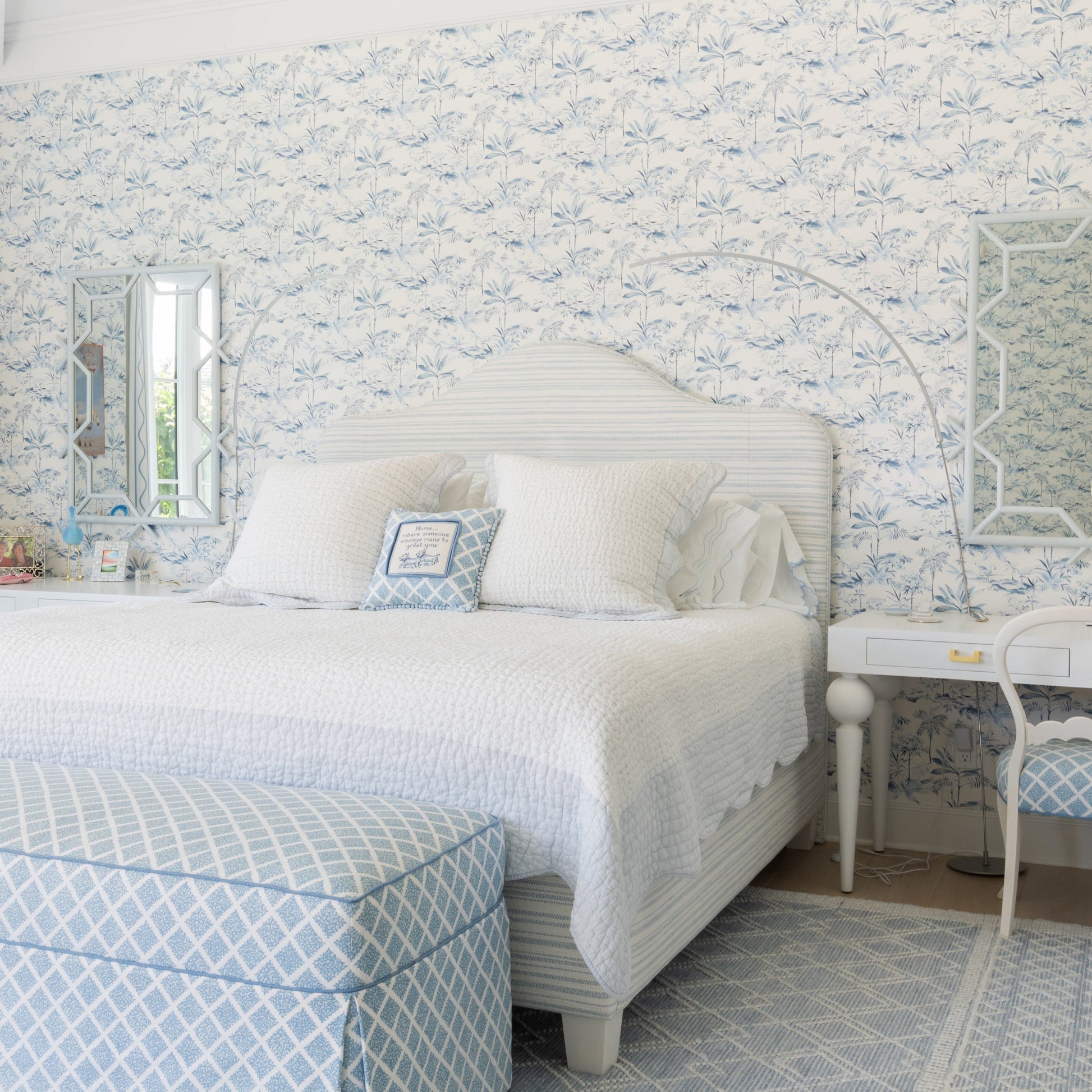 A beautifully styled bedroom featuring a white bed with white linens, a light blue and white patterned upholstered ottoman, a matching patterned chair, and a light blue and white geometric rug. The wall behind the bed is covered in matching blue and white floral toile wallpaper.