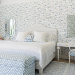 A beautifully styled bedroom featuring a white bed with white linens, a light blue and white patterned upholstered ottoman, a matching patterned chair, and a light blue and white geometric rug. The wall behind the bed is covered in matching blue and white floral toile wallpaper.
