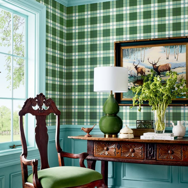 A styled interior corner with green-and-white plaid wallpaper, turquoise wainscoting, a dark carved wooden chair with a green cushion, a matching wooden console table, a green gourd lamp, and framed deer artwork.