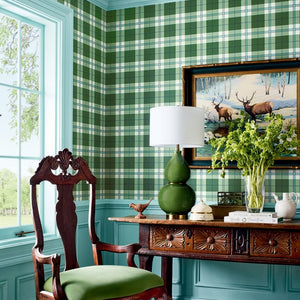 A styled interior corner with green-and-white plaid wallpaper, turquoise wainscoting, a dark carved wooden chair with a green cushion, a matching wooden console table, a green gourd lamp, and framed deer artwork.