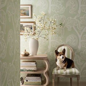 A dog sits on a striped upholstered chair in a styled interior room with pale green patterned wallpaper, a side table, books, and a vase of white blossoms.