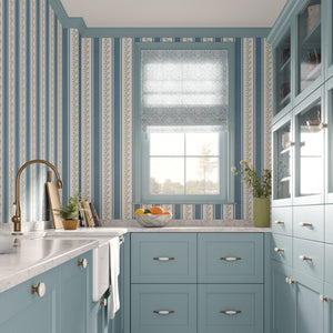 A beautifully styled kitchen featuring light blue cabinets, blue and white patterned wallpaper, a white marble countertop, a gold faucet, and a window with a sheer blue-gray roller shade.