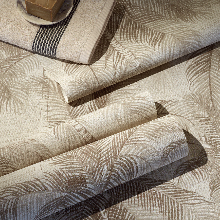 A close-up of folded wallpaper with a repeating palm-leaf pattern in warm beige and taupe tones, with a folded matching textile and a small soap dish with a bar of soap placed on top.